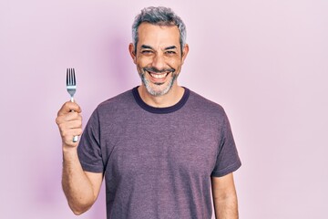 Handsome middle age man with grey hair holding one silver fork looking positive and happy standing and smiling with a confident smile showing teeth