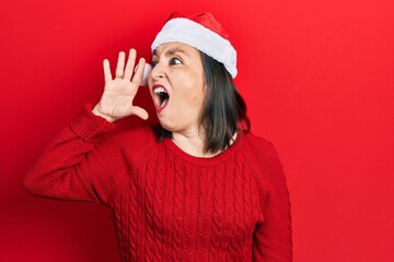 Middle age hispanic woman wearing christmas hat shouting and screaming loud to side with hand on mouth. communication concept.