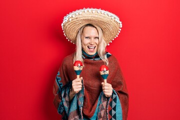 Beautiful caucasian blonde woman wearing festive mexican poncho and maracas smiling and laughing hard out loud because funny crazy joke.