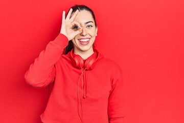 Beautiful woman with blue eyes wearing gym clothes and headphones smiling happy doing ok sign with hand on eye looking through fingers