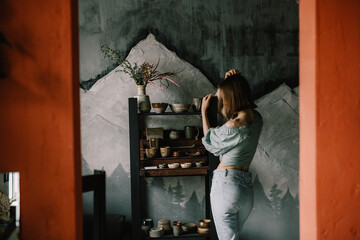 attractive girl in a blue blouse and light jeans is standing in a pottery workshop near a wooden shelf with pottery. Concept hobby, applied art