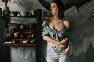 attractive girl in a blue blouse and light jeans is standing in a pottery workshop near a wooden...