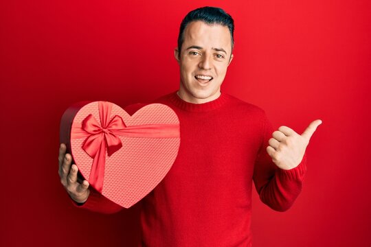 Handsome young man holding valentine gift pointing thumb up to the side smiling happy with open mouth