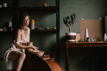 talented sculptor girl in thin light negligee sculpts a jug of clay in pottery workshop near large window. Concept art of sculpture