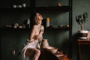 talented sculptor girl in thin light negligee sculpts a jug of clay in pottery workshop near large window. Concept art of sculpture