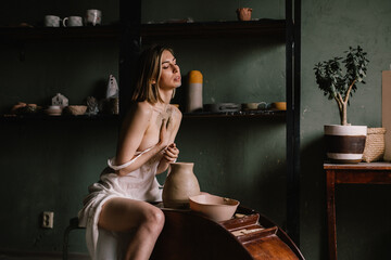 talented sculptor girl in thin light negligee sculpts a jug of clay in pottery workshop near large window. Concept art of sculpture