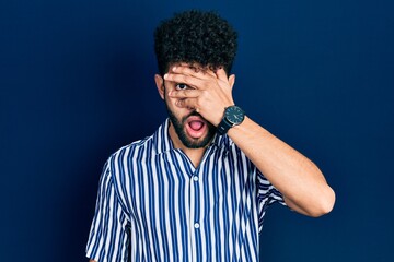 Young arab man with beard wearing casual striped shirt peeking in shock covering face and eyes with hand, looking through fingers afraid
