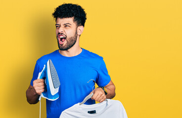 Young arab man with beard holding electric steam iron and white t shirt angry and mad screaming...