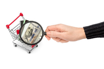 Woman research finances for shopping. Woman's hand holding magnifying glass over shopping cart with money. Isolated on white.