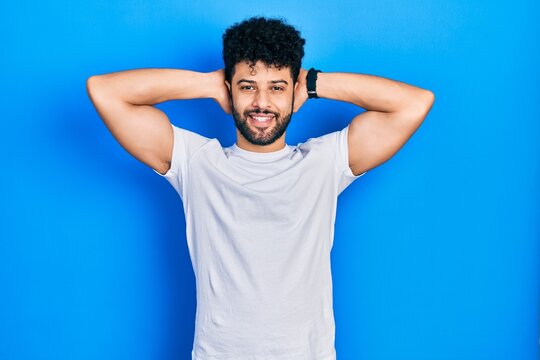 Young arab man with beard wearing casual white t shirt relaxing and stretching, arms and hands behind head and neck smiling happy