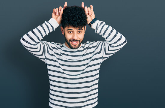 Young arab man with beard wearing casual striped sweater doing funny gesture with finger over head as bull horns