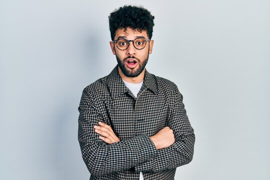 Young arab man with beard wearing glasses with arms crossed gesture afraid and shocked with surprise and amazed expression, fear and excited face.