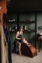 young sculptor, a girl with bandage on her head and in black apron, sculpts a clay product in pottery workshop near large window. Concept hobby sculpture
