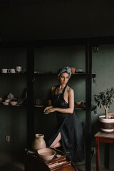 beautiful sculptor girl with bandage on her head and a black apron on her naked body is standing in pottery workshop near large window. concept is beauty and art.