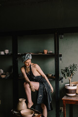 beautiful sculptor girl with bandage on her head and a black apron on her naked body is standing in pottery workshop near large window. concept is beauty and art.