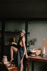 beautiful sculptor girl with bandage on her head and a black apron on her naked body is standing in pottery workshop near large window. concept is beauty and art.