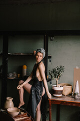 beautiful sculptor girl with bandage on her head and a black apron on her naked body is standing in pottery workshop near large window. concept is beauty and art.