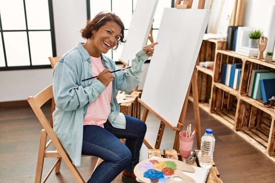 Middle age hispanic woman at art studio painting on canvas smiling happy pointing with hand and finger