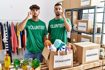 Young gay couple wearing volunteer t shirt at donations stand mouth and lips shut as zip with...