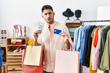 Young hispanic man holding shopping bags and credit card in shock face, looking skeptical and sarcastic, surprised with open mouth