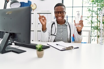 Young african doctor man holding hand sanitizer gel at the clinic smiling happy pointing with hand and finger to the side