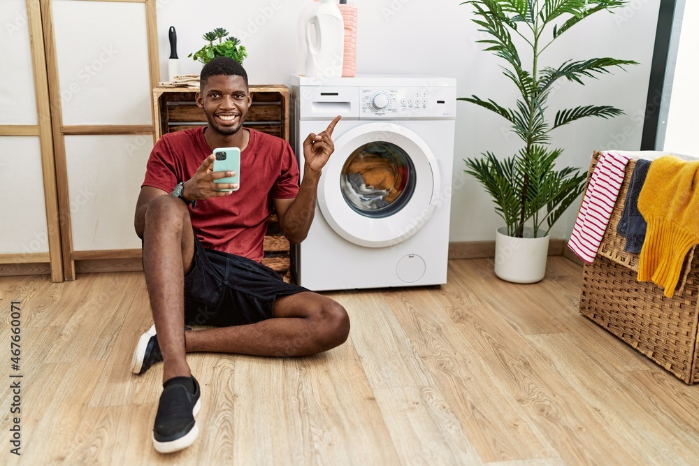 Poster Young african american man using smartphone waiting for washing machine cheerful with a smile on face pointing with hand and finger up to the side with happy and natural expression