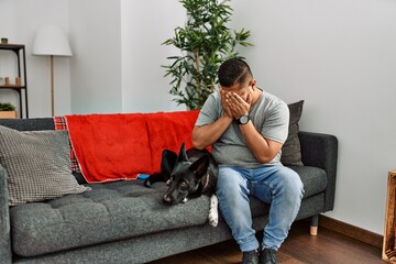 Young latin man and dog sitting on the sofa at home with sad expression covering face with hands while crying. depression concept.