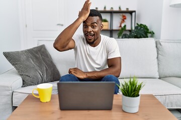 Young african man using laptop at home surprised with hand on head for mistake, remember error....