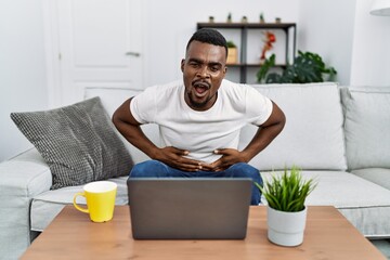 Young african man using laptop at home with hand on stomach because indigestion, painful illness feeling unwell. ache concept.