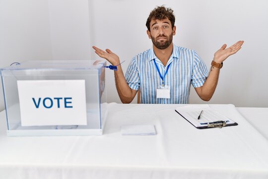 Young Handsome Man At Political Election Sitting By Ballot Clueless And Confused Expression With Arms And Hands Raised. Doubt Concept.