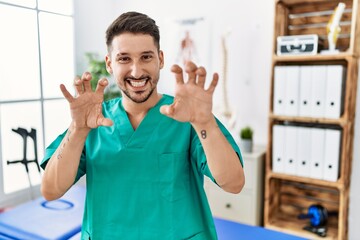Young physiotherapist man working at pain recovery clinic smiling funny doing claw gesture as cat,...