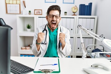 Young man with beard wearing doctor uniform and stethoscope at the clinic excited for success with arms raised and eyes closed celebrating victory smiling. winner concept.