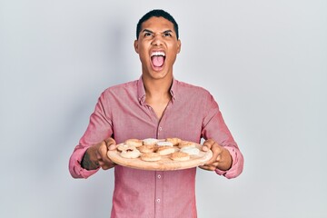 Young african american guy holding tray with cake sweets angry and mad screaming frustrated and furious, shouting with anger looking up.