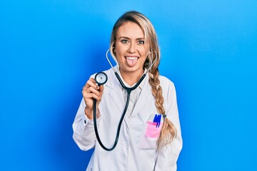 Beautiful young blonde doctor woman holding stethoscope sticking tongue out happy with funny expression. emotion concept.