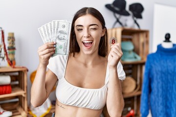 Young hispanic customer woman holding dollars banknotes and bitcoin winking looking at the camera with sexy expression, cheerful and happy face.