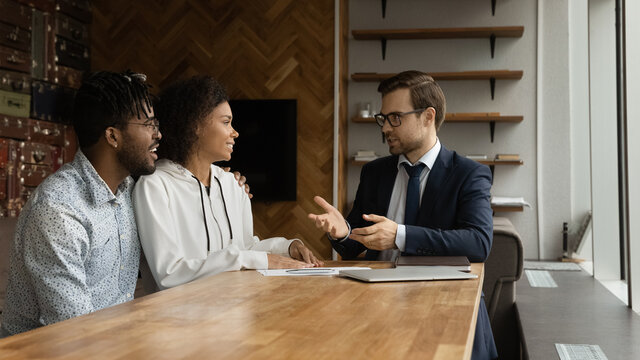 Positive smart young male real estate agent insurer lawyer giving professional consultation to happy family couple clients, making good propose or discussing good deal in modern office.