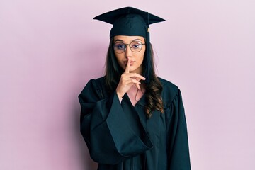 Young hispanic woman wearing graduation cap and ceremony robe asking to be quiet with finger on lips. silence and secret concept.