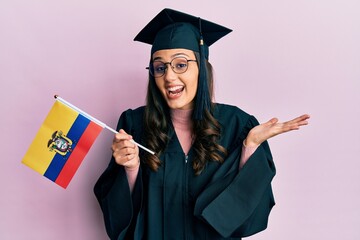 Young hispanic woman wearing graduation uniform holding ecuador flag celebrating achievement with happy smile and winner expression with raised hand