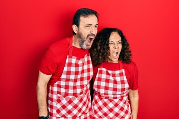 Middle age couple of hispanic woman and man wearing cook apron angry and mad screaming frustrated and furious, shouting with anger. rage and aggressive concept.