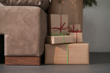 Presents in paper wrap with ribbons under christmas tree indoor near couch with warm light