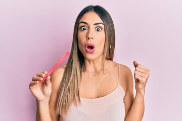 Young hispanic woman using file nail scared and amazed with open mouth for surprise, disbelief face