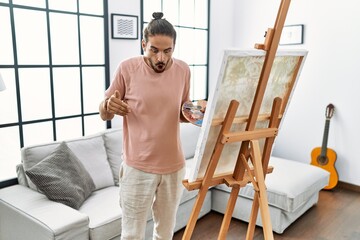 Young hispanic man with beard painting on canvas at home pointing down with fingers showing advertisement, surprised face and open mouth