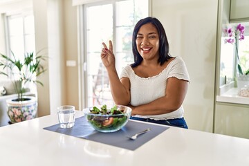 Obraz na płótnie Canvas Young hispanic woman eating healthy salad at home smiling with happy face winking at the camera doing victory sign. number two.