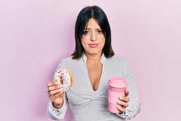 Young hispanic woman eating doughnut and drinking coffee skeptic and nervous, frowning upset...