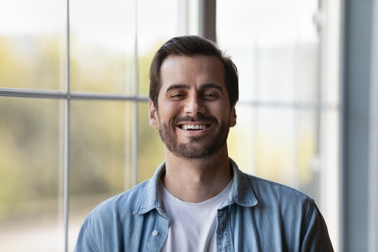 In a perfect mood. Headshot portrait of young man standing indoors near large window laugh demonstrate white healthy teeth. Profile picture of happy casual millennial guy look at camera feel motivated