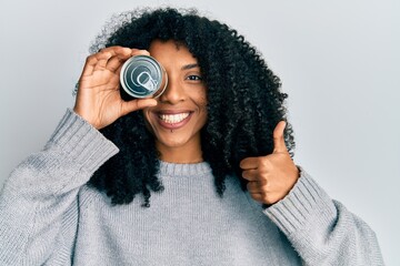 African american woman with afro hair holding canned food on eye smiling happy and positive, thumb...