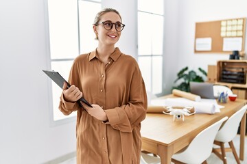 Young blonde woman smiling confident holding clipboard at architect studio