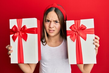 Beautiful brunette little girl holding gifts clueless and confused expression. doubt concept.