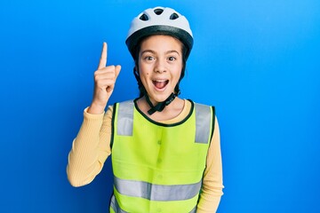 Beautiful brunette little girl wearing bike helmet and reflective vest pointing finger up with successful idea. exited and happy. number one.