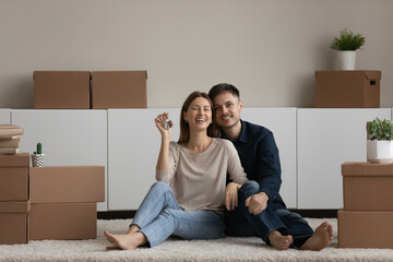Portrait happy couple demonstrating showing keys, looking at camera, sitting on floor with cardboard boxes in new home, enjoying relocation, smiling woman and man hugging, celebrating moving day
